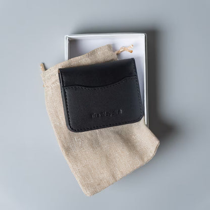 A black leather card holder wallet with debossed 'The Bicyclist' branding, resting on a beige linen drawstring pouch inside an open white box, set against a solid gray background.