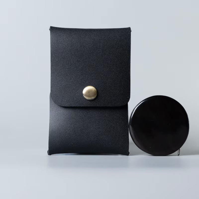 A black full grain leather card pouch or card holder with a gold snap button closure standing upright, made by 'The Bicyclist' next to a round black shelac box on a light gray studio background.