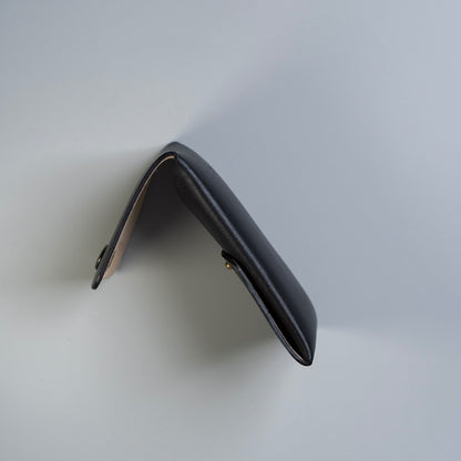 A black leather card holder or card wallet shown from an angled top-down view, featuring a small gold-tone snap button closure and a light beige felt interior lining, made by, 'The Bicyclist' set against a clean light gray background.
