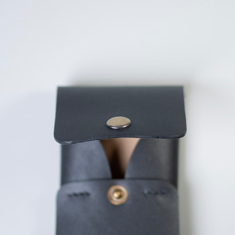 Close-up of a black full grain leather flap with a snap-button closure and a brass-toned rivet, of a leather card holder or a card pouch made by 'The Bicyclist' photographed against a plain light background.