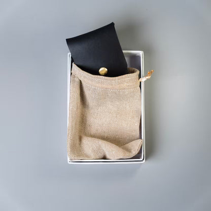 A black full grain leather card holder or a card pouch with a gold snap button, made by 'The Bicyclist' half tucked inside a beige burlap drawstring pouch, placed inside a white rectangular box on a neutral gray background.
