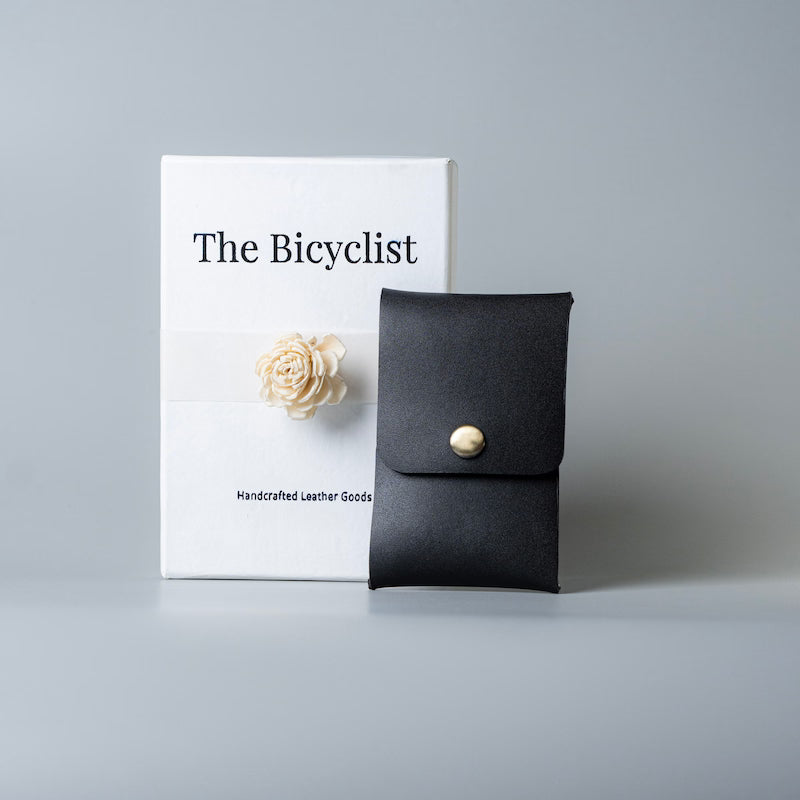A black full grain leather card pouch or card holder with a gold snap button, standing next to its white packaging box labeled 'The Bicyclist' on a light gray background.