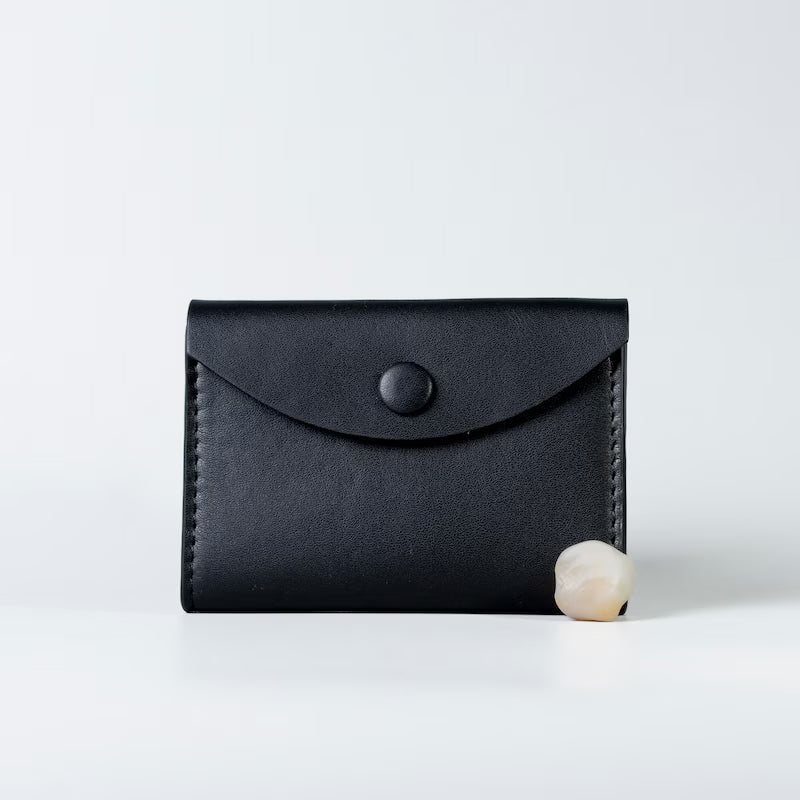A black full grain leather card holder wallet with a flap closure and snap button from 'The Bicyclist', shown on a clean white background with a small pearl prop at its base.