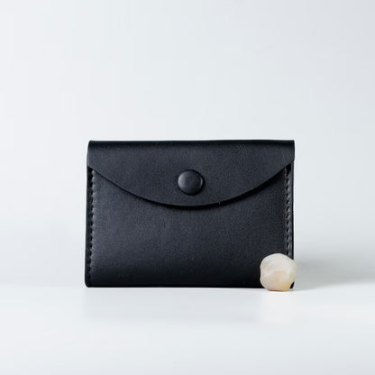 A black full grain leather card holder wallet with a flap closure and snap button from 'The Bicyclist', shown on a clean white background with a small pearl prop at its base.