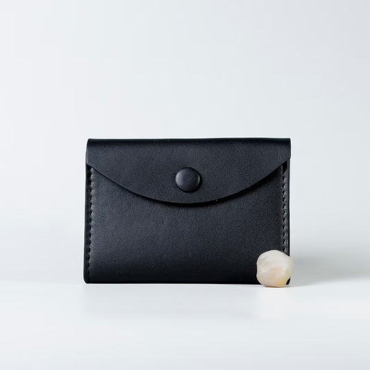A black full grain leather card holder wallet with a flap closure and snap button from 'The Bicyclist', shown on a clean white background with a small pearl prop at its base.