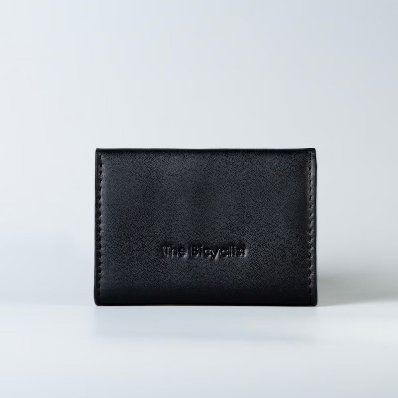 A black full grain leather card holder wallet with 'The Bicyclist' debossed on the back, set against a clean white studio background.