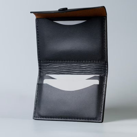 A black full grain leather card holder wallet shown open, displaying its interior compartments, including a clear ID window and a tan sheep leather lining on the inside flap made by 'The Bicyclist' set against a light gray background.