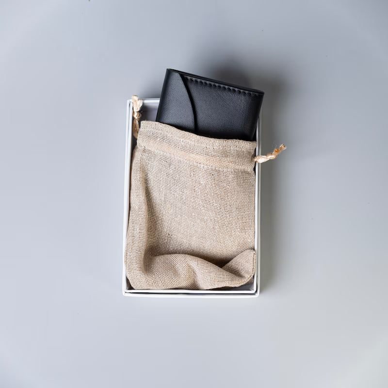 A black full grain leather wallet tucked inside a white rectangular box with a beige burlap drawstring pouch, photographed from above on a light gray background.
