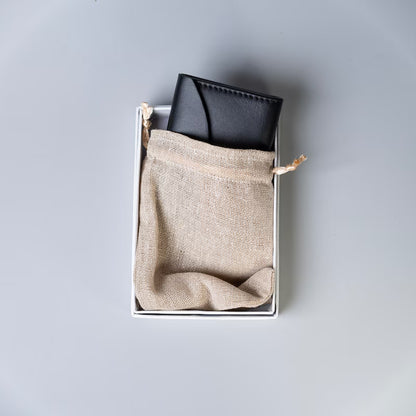 A black full grain leather wallet tucked inside a white rectangular box with a beige burlap drawstring pouch, photographed from above on a light gray background.