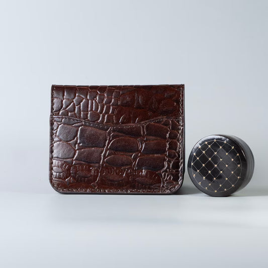 Full grain leather maroon slim bifold card wallet in corocodile leather texture with a card slot from The Bicyclist next to a shelac box on a soft grey background 