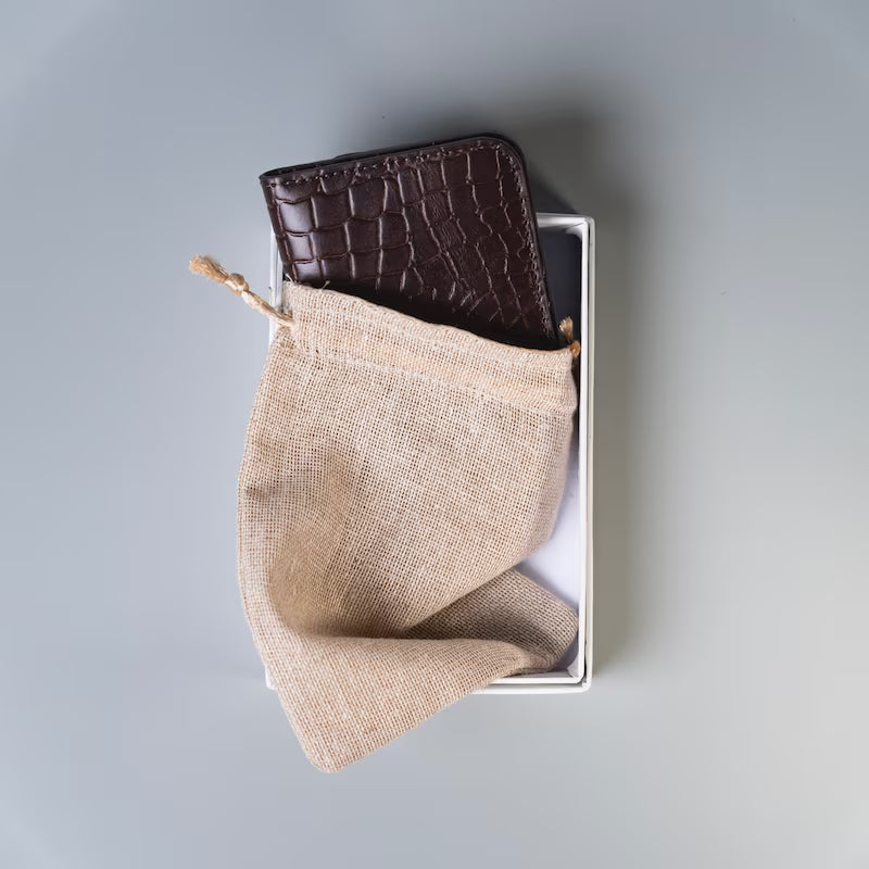 A bifold  full grain leather card wallet in crocodile texture leather half inserted into a beige cloth pouch placed inside a  paper cardboard gift box on a grey background