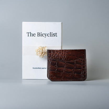Maroon Full grain leather bifold card wallet with crocodile leather texture placed upright in front of a gift box with  the brand name, The Bicyclist printed on it with a off white paper flower 