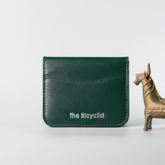 A dark green full grain leather card holder wallet with white embossed text reading 'The Bicyclist' on the front, displayed on a light gray background next to a small gold horse figurine.