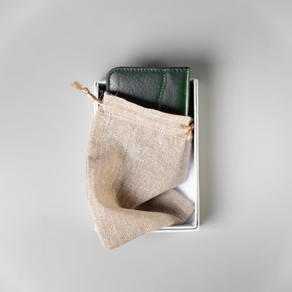 A dark green full grain leather card wallet made by, 'The Bicyclist' tucked half inside a jute drawstring dust pouch kept inside a white cardboard box on a soft grey background