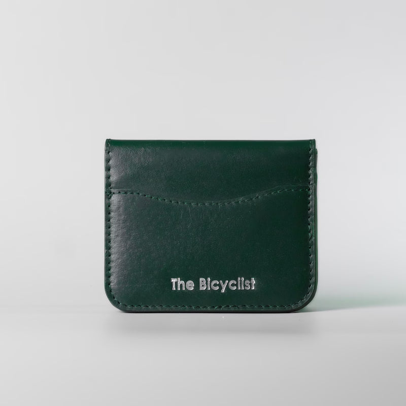 A dark green full grain leather card holder wallet with white embossed text reading 'The Bicyclist' on the front, set against a light gray background.
