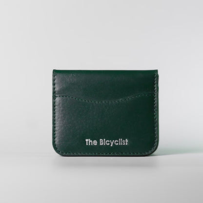 A dark green full grain leather card holder wallet with white embossed text reading 'The Bicyclist' on the front, set against a light gray background.