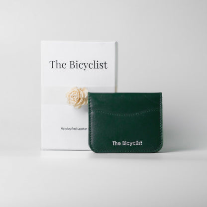 A dark green leather card holder wallet with 'The Bicyclist' logo, displayed in front of its white packaging box which features the same brand name and a small cream-colored flower.