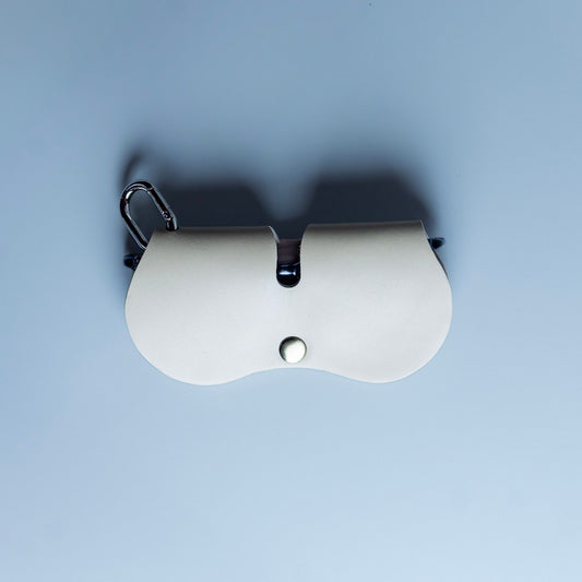 A off white leather spectacle and eyeware protective sleeve with a snap closure, from The Bicyclist featuring a chrome carabiner attached to the top right, set against a solid light blue background.