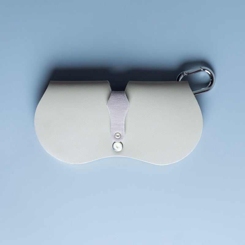 Back view of an off white leather spectacle and eyeware protective sleeve  from The Bicyclist with a central strap and snap closure, featuring a chrome carabiner attached to the top right, set against a solid light blue background.