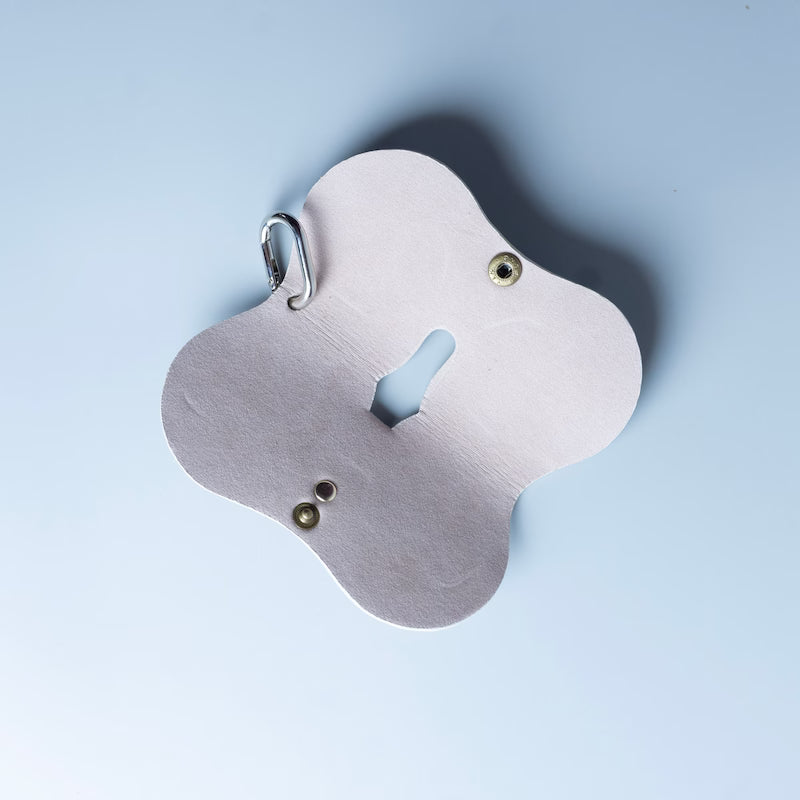 Full open view of a off white leather spectacle and eyeware protective sleeve made by 'The Bicyclist', featuring a chrome carabiner attached to the top right, set against a solid light blue background.