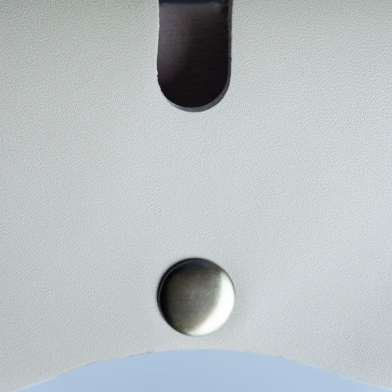 Close-up detail of a off white leather surface with a dark oval cutout near the top and a round metallic rivet near the bottom.
