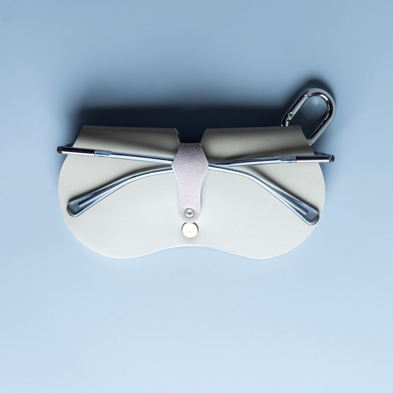 A pair of silver metal-framed eyeglasses resting inside a light beige leather case with a matching carabiner hook, made by 'The Bicyclist' set against a solid light grey background.