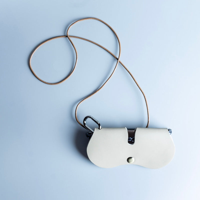 A cream-colored leather eyeware and sunglasses sleeve with a curved silhouette, attached with a thin brown leather neck strap, and a silver metal snap closure, photographed on a light blue background.