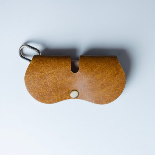 A brown leather spectacle and sunglasses case with a metal carabiner clip and a snap button closure, made by, 'The Bicyclist' photographed from above on a light gray background.