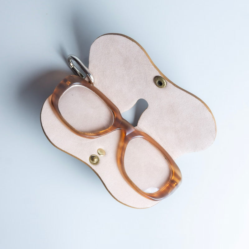 A pair of translucent amber acetate eyeglasses displayed on a light beige, butterfly-shaped leather spectacle sleeve handmade by 'The Bicyclist' with brass snap buttons and a silver metal clip, shot from a top-down angle on a pale blue background.