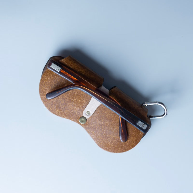 Rear view of a pair of amber acetate eyeglasses inside a brown textured leather case with a silver metal carabiner clip made by 'The Bicyclist', shot from a top-down angle on a light grey background.