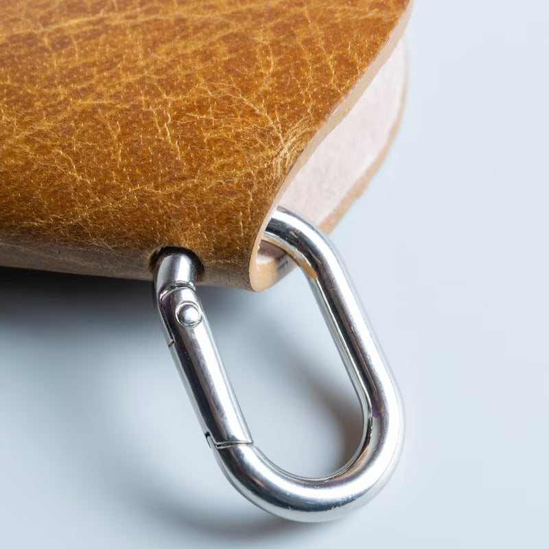 Close-up of a brown leather spectacle sleeve with a silver metal carabiner clip attached, photographed on a light gray background.
