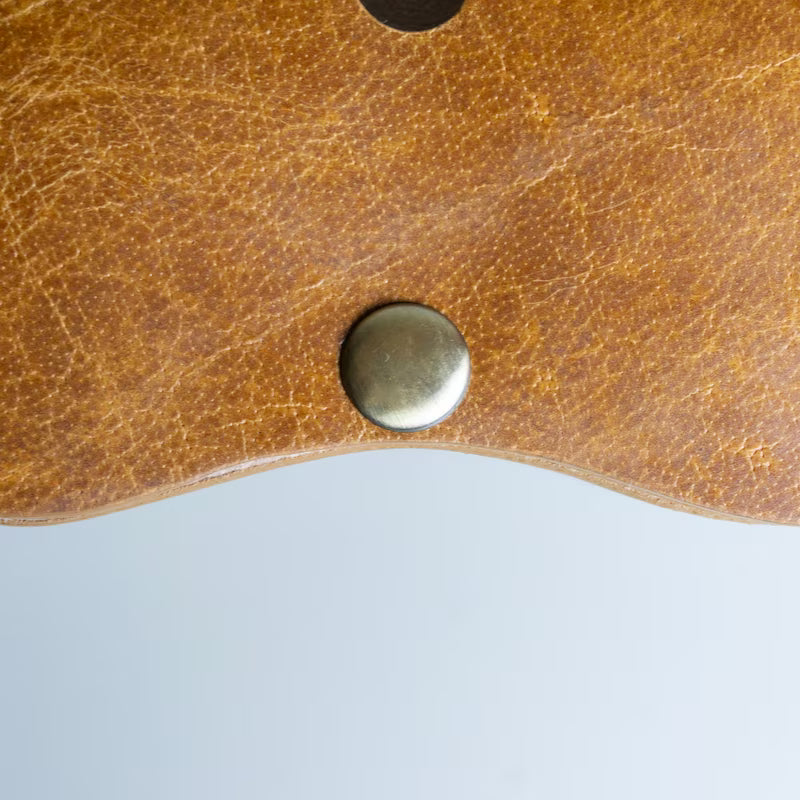 Close-up of a textured brown leather sunglasses sleeve with a round metal snap button, set against a light gray background.