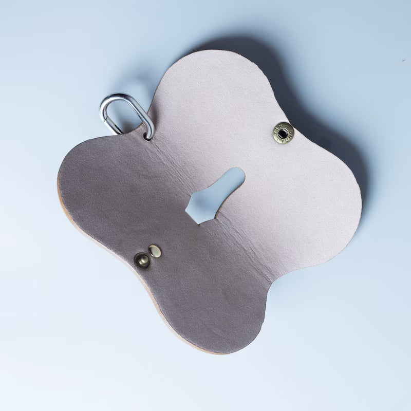 A clover-shaped leather spectacle sleeve with a dark gray exterior, a light beige interior, a hole cutout, and a silver metal 'D' ring, shown on a light blue background.