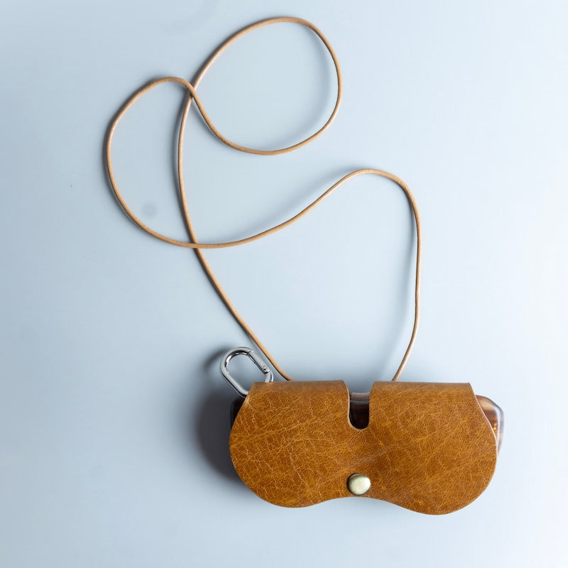 A tan leather spectacle and sunglasses sleeve with a matching adjustable neck strap, featuring a silver metal carabiner clip and a small white snap button,  made by The Bicyclist, shown on a light grey background.