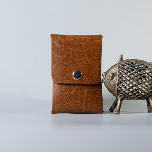 A brown textured full grain leather card pouch with a snap button closure made by 'The Bicyclist', displayed next to a decorative metal fish figurine on a light gray background.