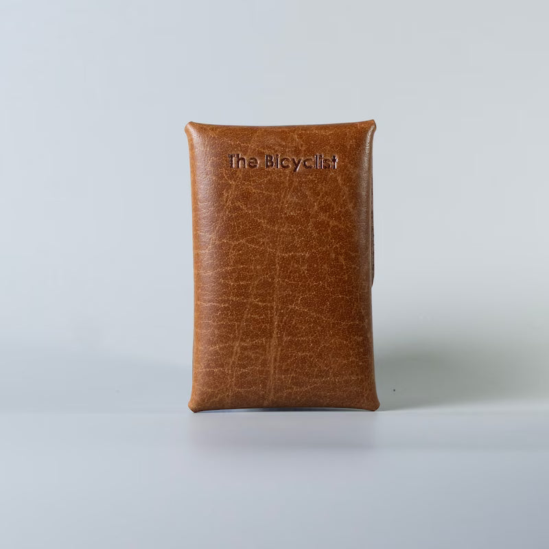 A tan brown full grain leather pouch with a textured finish, featuring the debossed brand name 'The Bicyclist' at the top, set against a light gray background.