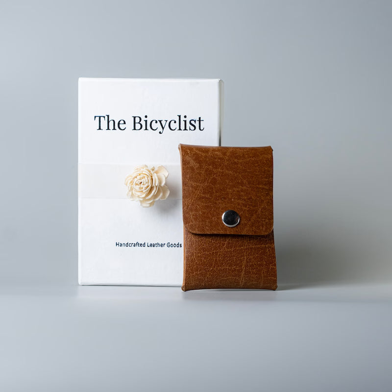 A tan brown leather card holder or card pouch with a snap button closure stands in front of a white box labeled 'The Bicyclist' on a light gray studio background.