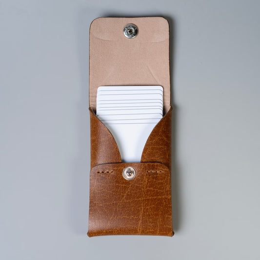 A tan brown full grain leather card holder or card wallet with a flap and a silver snap button, shown open with white plastic cards partially inserted, made by 'The Bicyclist' on a neutral gray background.