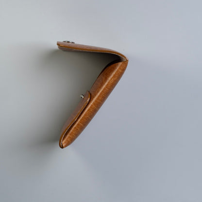 Side view of a tan brown leather card holder or card wallet with a curved, folded shape, shown on a plain light gray background.