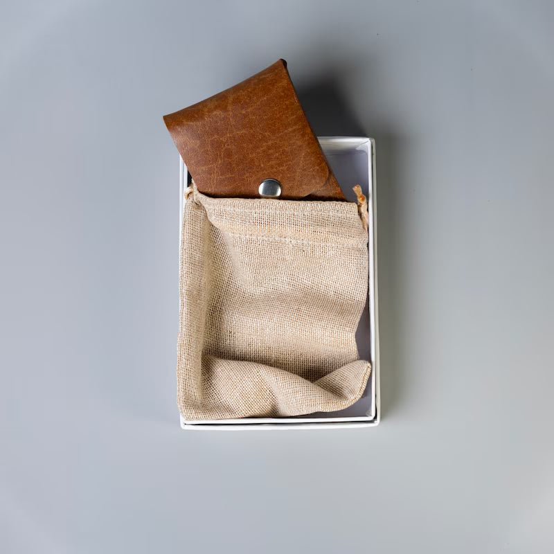 A tan brown leather card holder with a silver snap button, made by 'The Bicyclist' shown partially inserted into a beige drawstring jute dust pouch in a white carboard gift box, on a neutral gray background.