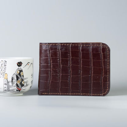 A maroon crocodile textured bifold full grain leather wallet next to a hand painted Japanese Tea  cup on a grey background
