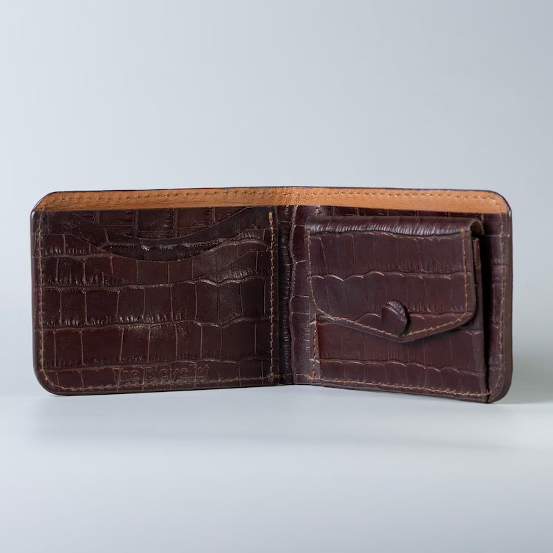 A maroon crocodile-embossed full grain leather bifold wallet from The Bicyclist shown open, revealing card slots and an interior pocket, set against a light gray background.