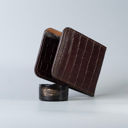A maroon crocodile-embossed full grain leather bifold wallet standing upright, with a black shellac accessory resting on a small round black stand against a light gray studio background.