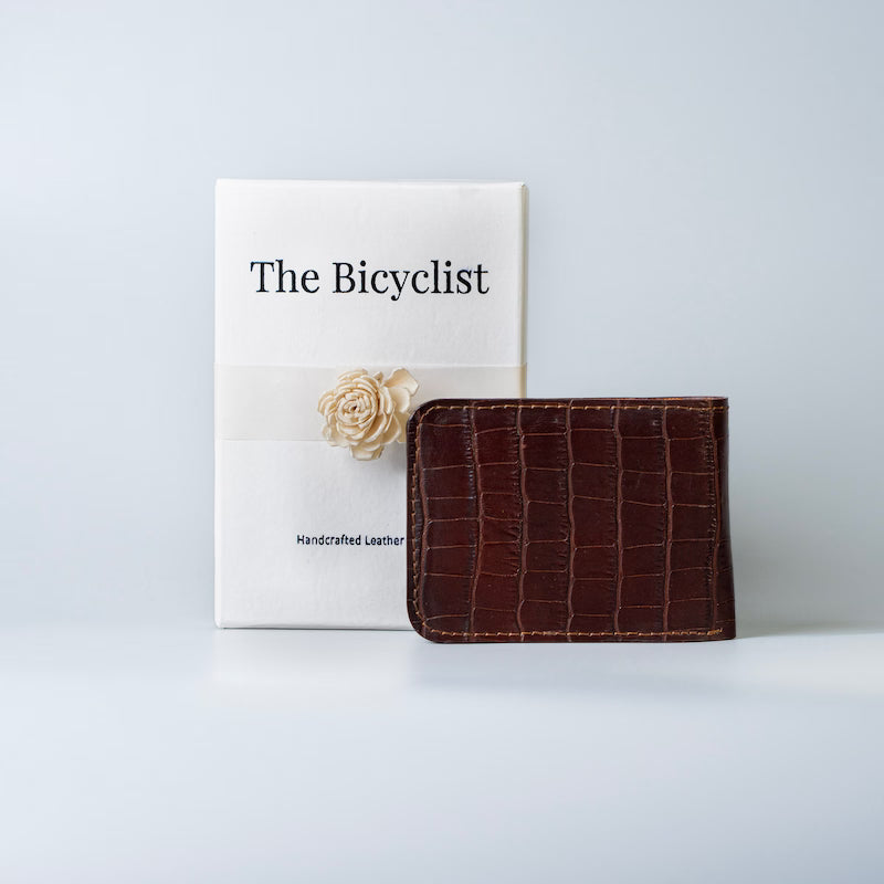 A maroon crocodile-embossed full grain leather bifold wallet displayed next to its white packaging box, which features the text 'The Bicyclist' and a cream-colored decorative flower.