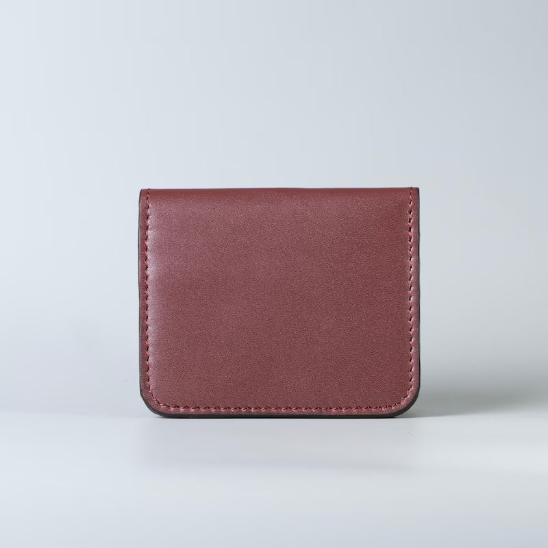 A slim compact maroon full grain leather card holder wallet with visible stitching from The Bicyclist, photographed upright against a light gray studio background.