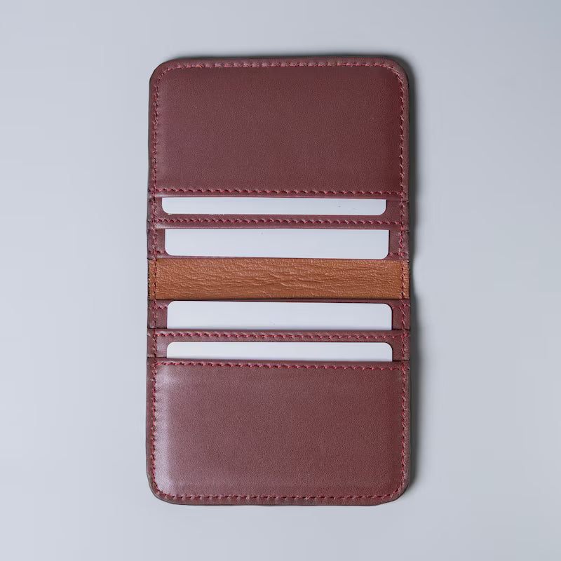 A maroon full grain leather card holder wallet shown open with white plastic cards inserted in the card slots from The Bicyclist on a light gray background, featuring multiple card slots and a contrasting tan leather lining.
