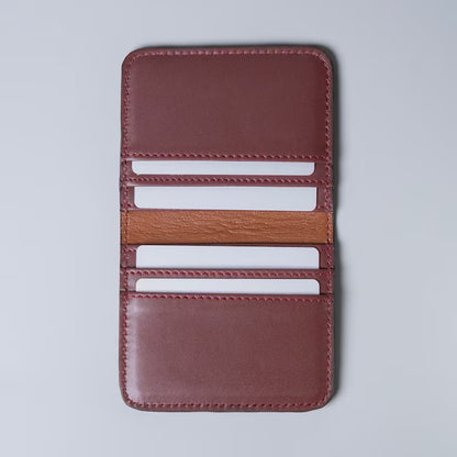 A maroon full grain leather card holder wallet shown open with white plastic cards inserted in the card slots from The Bicyclist on a light gray background, featuring multiple card slots and a contrasting tan leather lining.