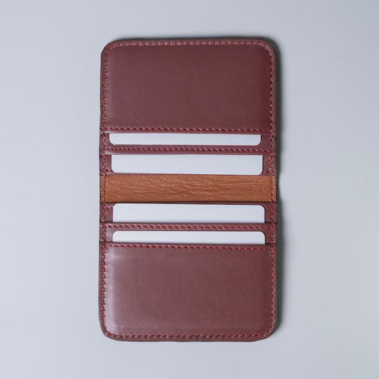 A maroon full grain leather card holder wallet shown open with white plastic cards inserted in the card slots from The Bicyclist on a light gray background, featuring multiple card slots and a contrasting tan leather lining.
