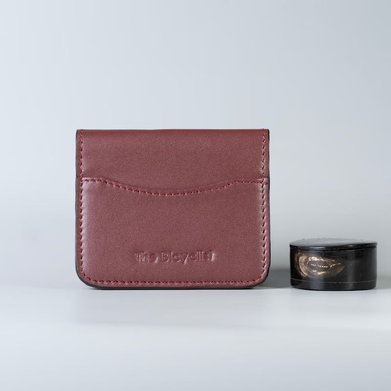 A maroon full grain leather card holder wallet with 'The Bicyclist' embossed on the front, shown next to a small black cosmetic shelac case on a light gray background.