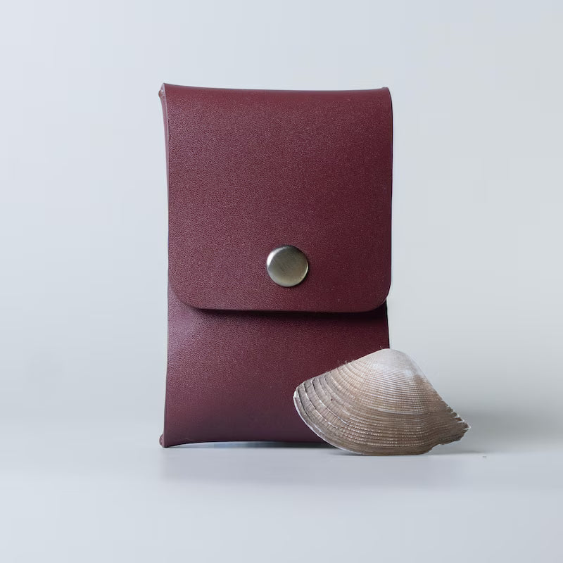 A burgundy leather pouch with a snap button closure, made by 'The Bicyclist' shown standing upright next to a small clam shell on a light gray background.